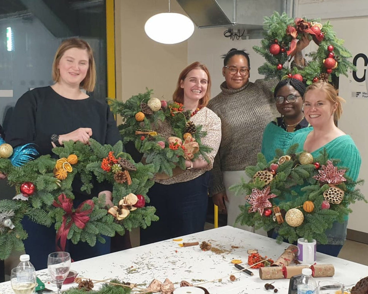 A group of six people standing together in an indoor studio or workshop, each holding a handmade Christmas wreath made of green pine branches with colorful ornaments and floral decorations. A white table covered with craft supplies and pinecones sits in the foreground, with a ring light, office chair, and white brick wall in the background.